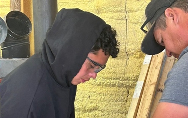 A young man in a hoodie and safety glasses works alongside an instructor during a Society of Indian Missions knife-making class